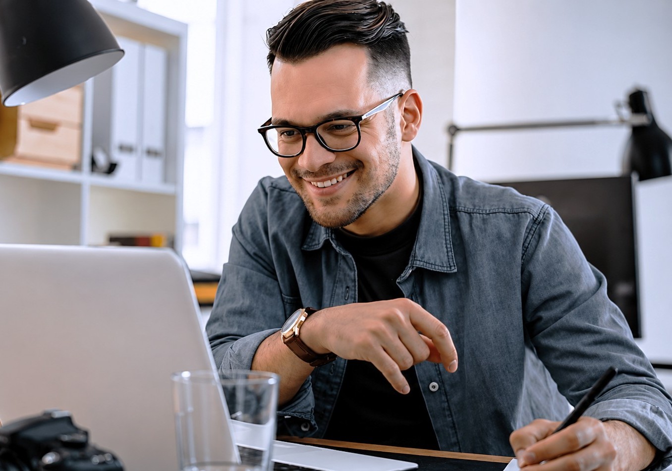 Smiling professional in glasses writing at a desk with a laptop in a bright office